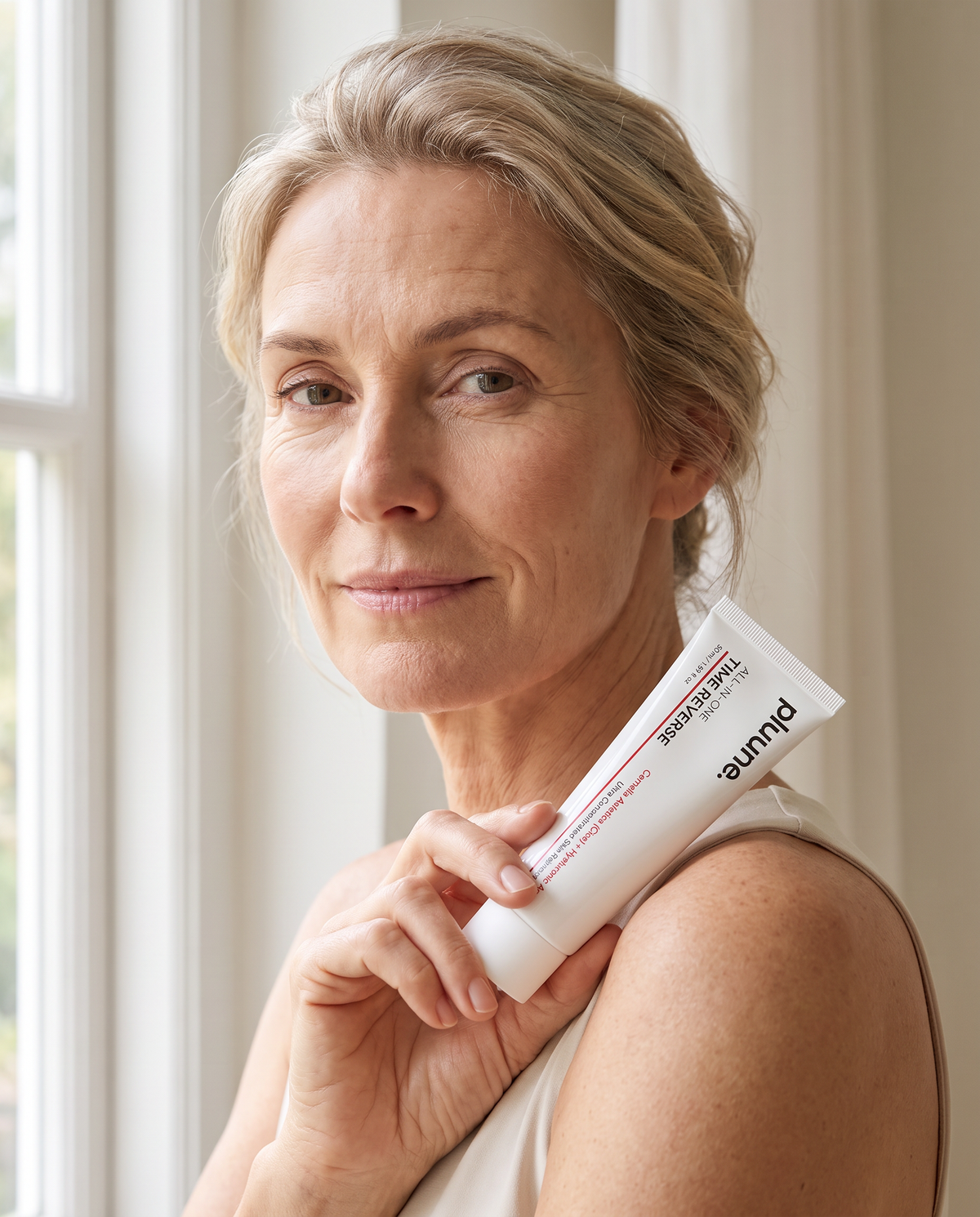 Woman with blonde hair pulled back holding a tube of Pluune Time Reverse cream against her shoulder by a window