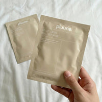 Hand holding a Pluune collagen sheet mask sachet over white bedding, a second sachet lying beside it
