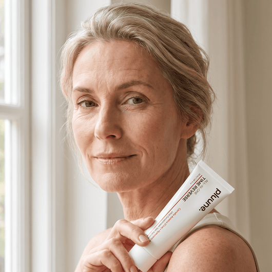 Woman with gray-blonde hair by a window holding a Pluune Time Reverse Cream tube against her shoulder